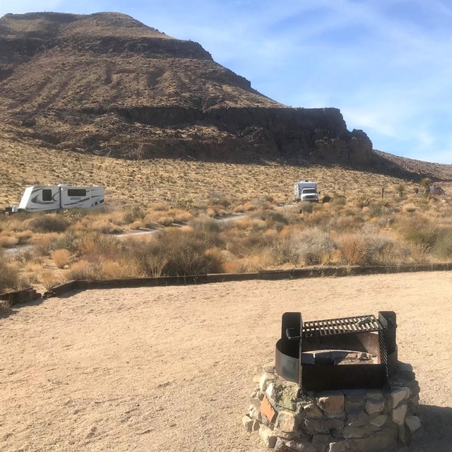 A bluff in the background with RVs. A campfire pit and picnic table in the foreground. 