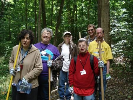 Six men and women in work gloves stand holding shovels in a forest.