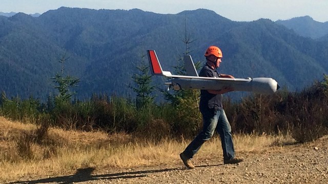 A man in a helmet carries a UAS on a road.