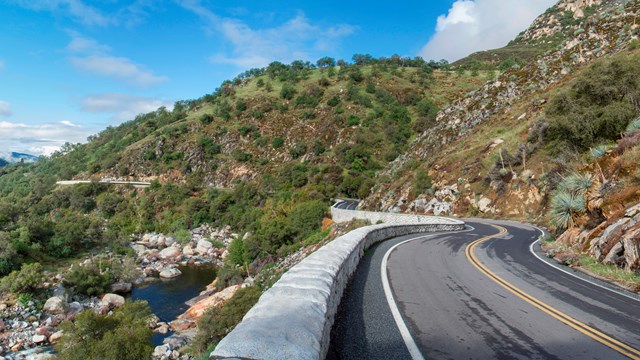 A stretch of road that curves around a bend. The road is surrounded by vegetation and a river.