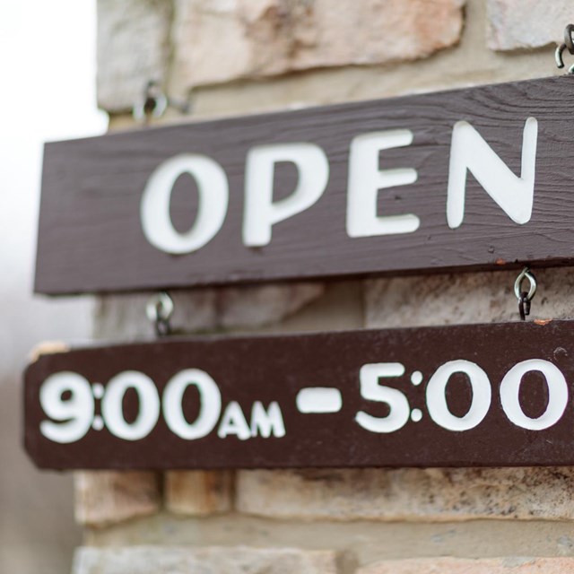 A brown open sign hanging from a stone wall. A small sign under it reads: 9 a.m. to 5 p.m.