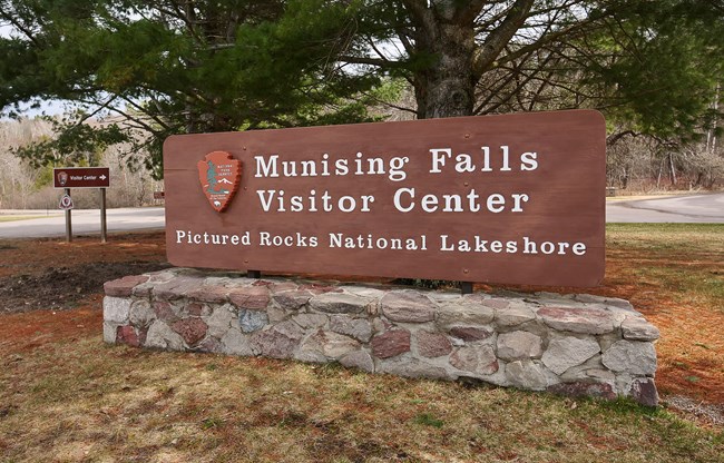 A brown sign with a stone base that reads "Munising Falls Visitor center"