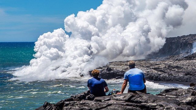 Two hikers sitting watching a volcano next to the ocean erupt 