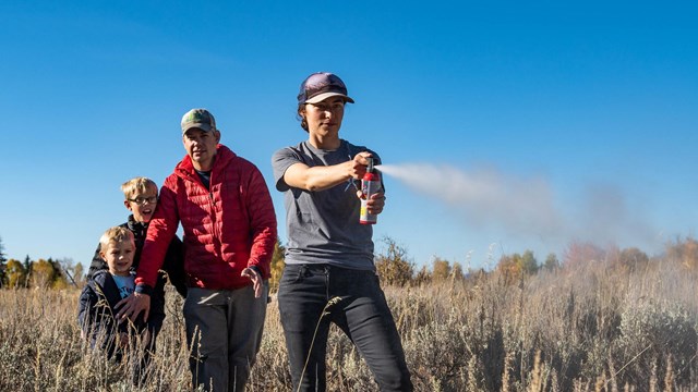 Woman spraying bear spray while man with boy stands in sagebrush behind.