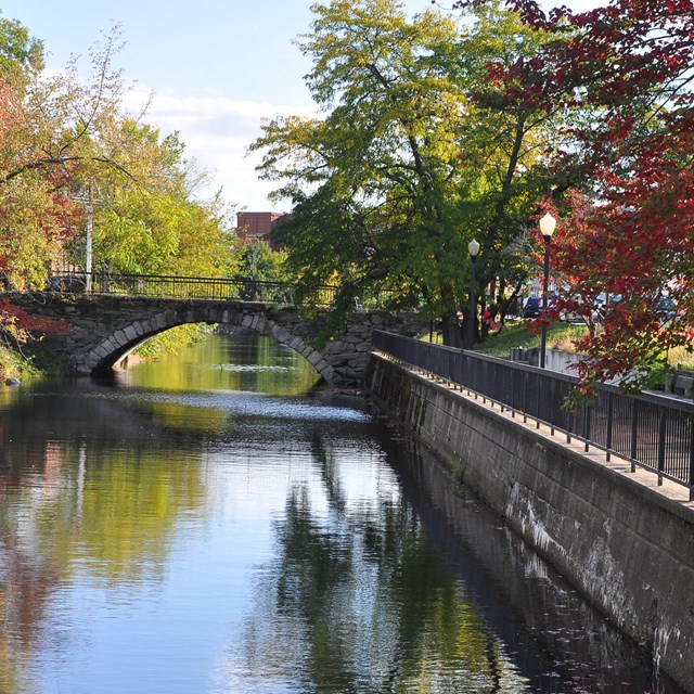 a bridge over canal on left and walkway with autumn trees and grass on right