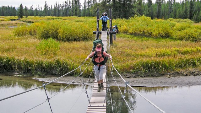 Hikers crossing a creek via a cable suspension bridge.