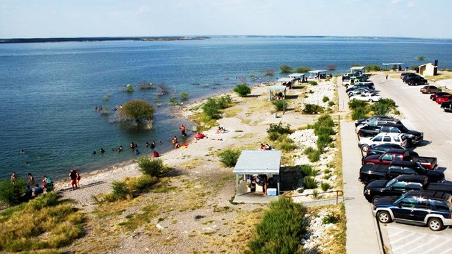 7 ramadas shading picnic tables bordered on right by parking lot with a vault toilet station and on left by lake. People are picnicking, sitting on sandy lakeshore, and swimming.