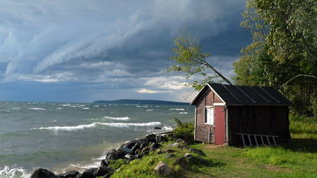 Storm clouds are visible in the distance as waves crash against a rocky shoreline near a small shed.