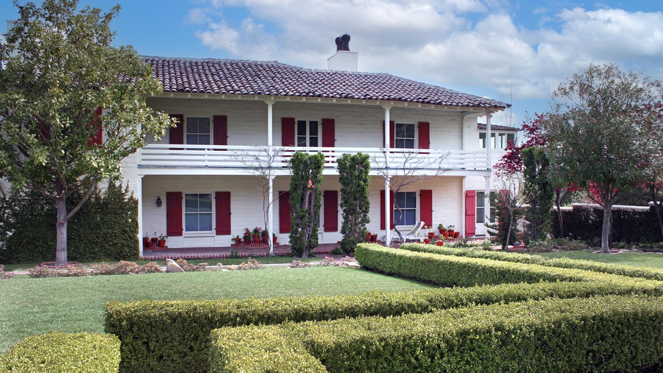 Hedges, trees and a courtyard are seen next to a two story home with shutters.