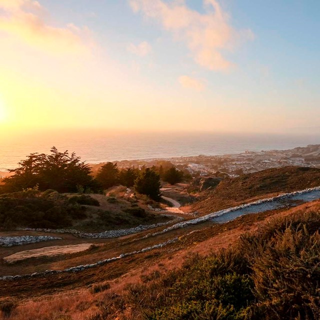 sunsets on dirt road that curves down hillside above ocean front houses