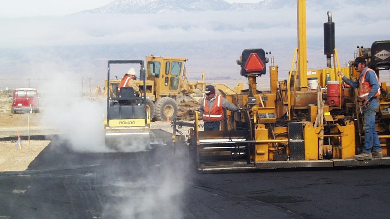 Various large yellow industrial machines sit atop a steaming black patch of asphalt, likely pavingit