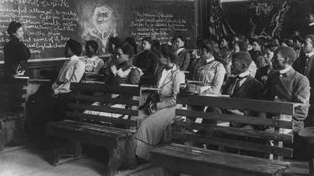  B&W photo of teacher standing in front of classroom of students with writing on blackboard