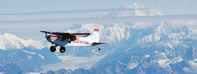 Fixed wing single engine aircraft flying under blue skies and above snowy mountains.