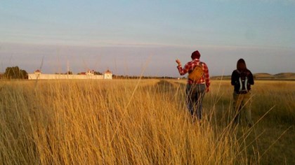 Two people walk in a field towards a white walled Fort. 