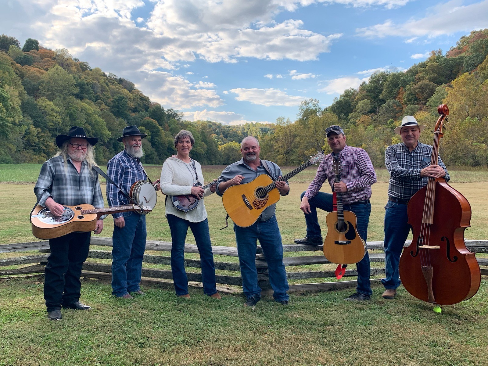 One Lane Bridge Six musicians with instruments standing in front of a split rail fence