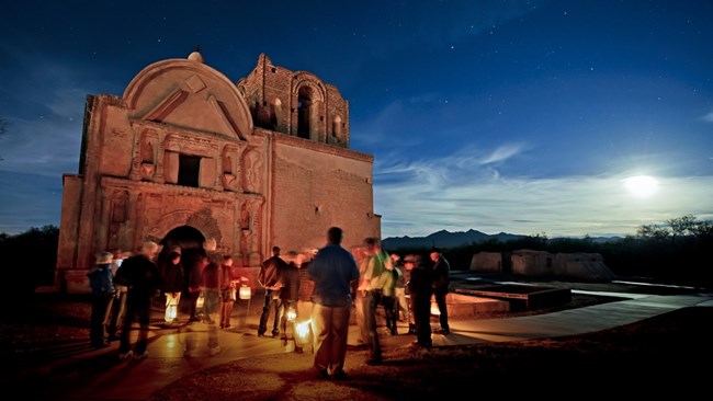 Evening Program at Tumacácori National Historical Park visitors carrying lanterns gather around the ruins of an adobe brick mission church at night