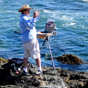 Photo of an artist with an easel along a rocky shoreline using a phone to snap a photo of the scene he is painting