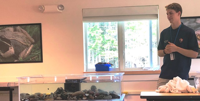 A young man stands to the right, there is a table to the right of him with water, rocks, and seaweed in it.