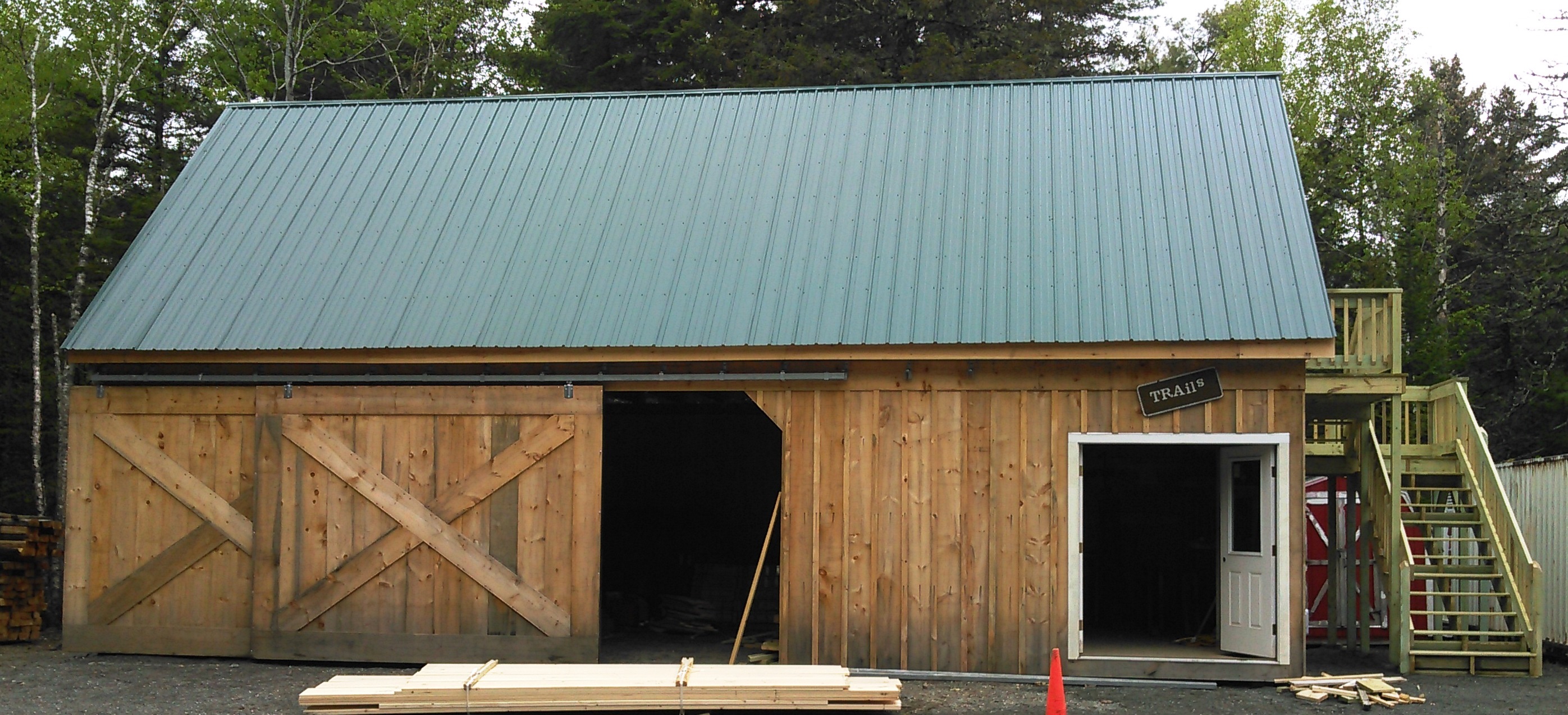 A pine wood, two story shed with barn doors open to the left and regular doors open to the right stands facing the viewer. A green metal roof covers the structure, while small amounts of cloudy sky can be seen surrounding the structure.