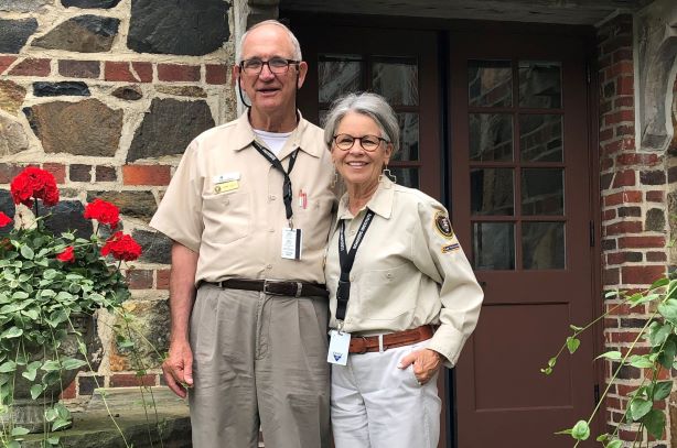 Acadia Volunteers Tommy and Sandra Jarrett, pose together in their uniforms, facing the camera and smiling, while standing on stone steps, with red geraniums in blooms around them.