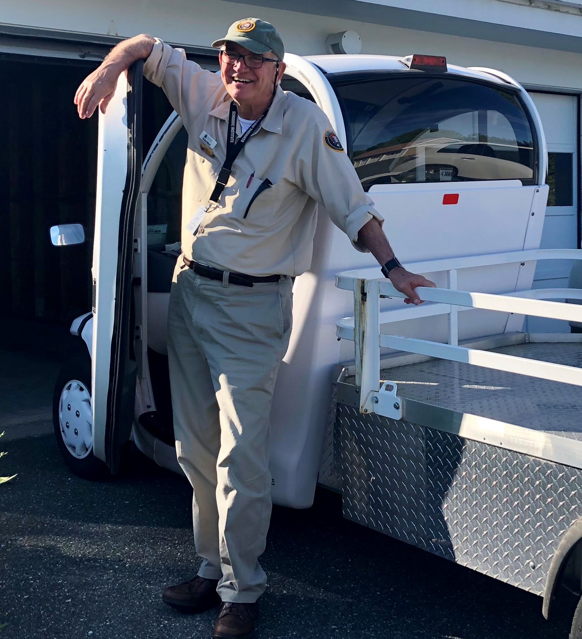 Volunteer Tommy Jarrett smiles at the camera as he stands leaning against the open doors of a small white electric vehicle. He is in his khaki colored uniform, is wearing a ball cap and glasses.