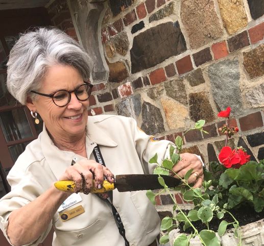 Volunteer Sandra Jarrett holds a muddy trowel above a flower box with w bright red blooming geranium.