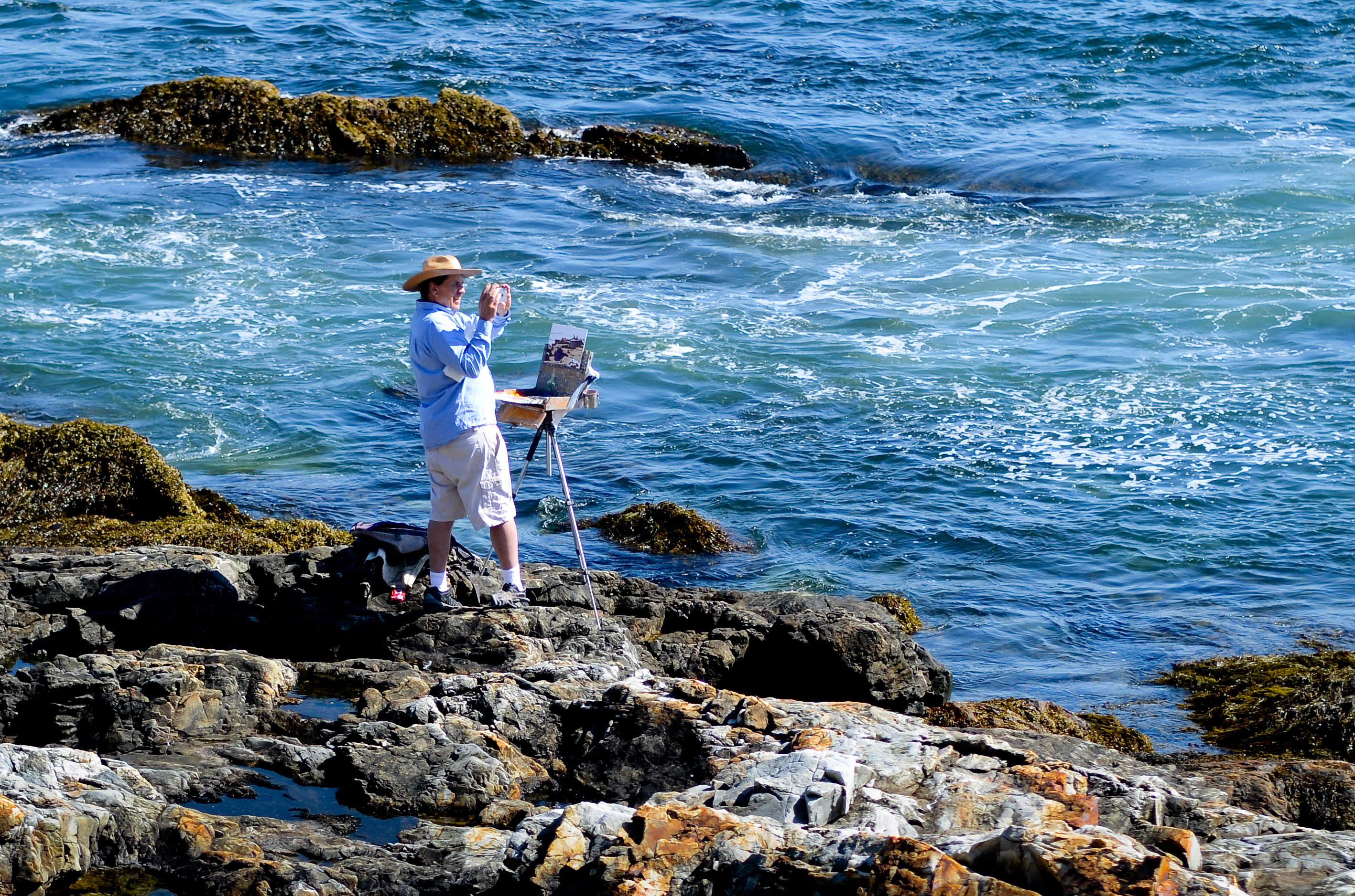 Artist standing on rocks takes a snapshot of the coastline he is painting