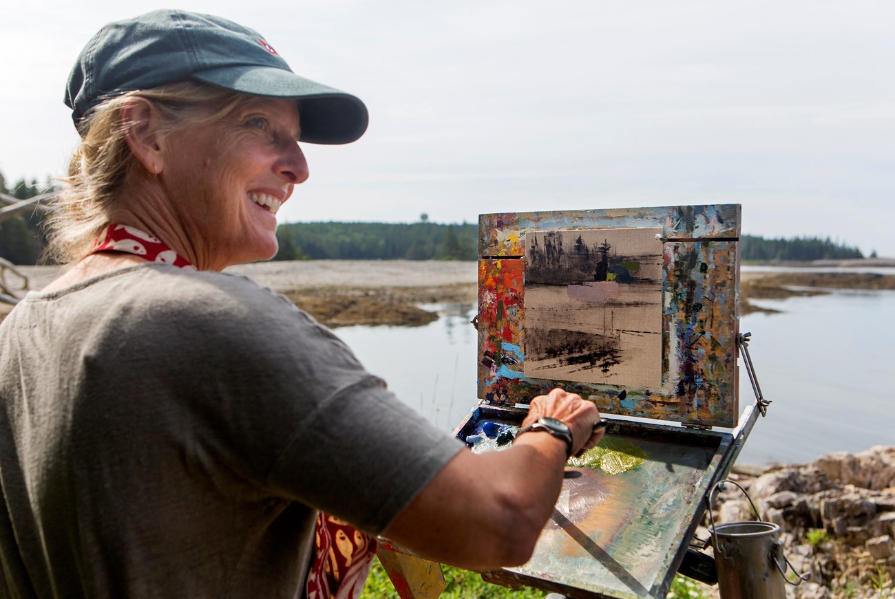 Woman in ball cap smiles while painting on an easel perched along coastline