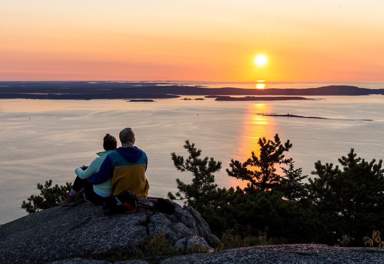two people sit on summit staring at sun rising above the ocean