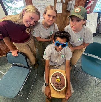 Three young women stand above a young man wearing Happy Birthday glasses with a cake on his lap