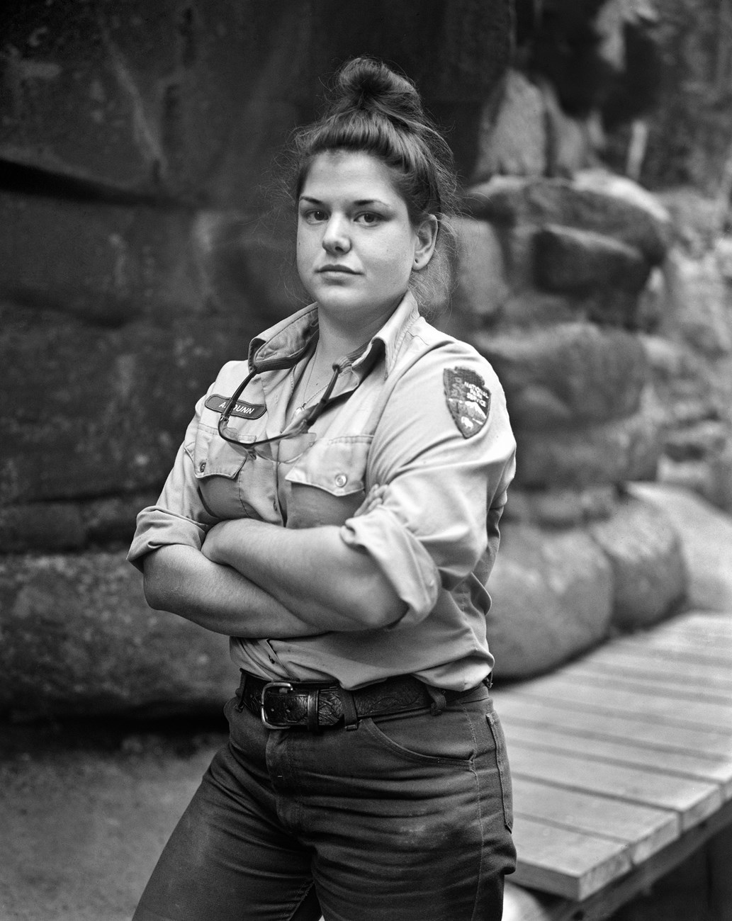 Black and white photograph of a trail crew worker with their arms crossed. Forest scene in the background.