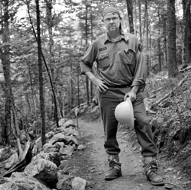 Black and white photograph of a trail crew worker standing in the middle of a trail with his hat in his hands. He is wearing a piece of cloth tied around his head.