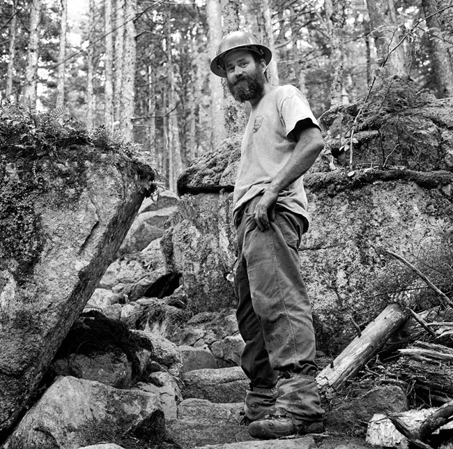 Black and white photograph of a male trail crew worker standing next to large rocks and wearing a hard hat. He has a large beard.