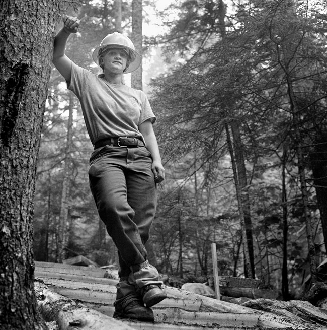 Black and white photograph of a female trail crew worker in the forest, wearing her hard hat, and using her arm to lean against a tree.