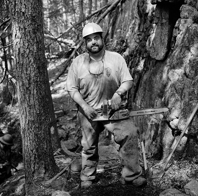 Black and white photograph of a male trail crew worker in the forest and wearing a hard hat. He is leaning against one leg and has a chainsaw in his hand.