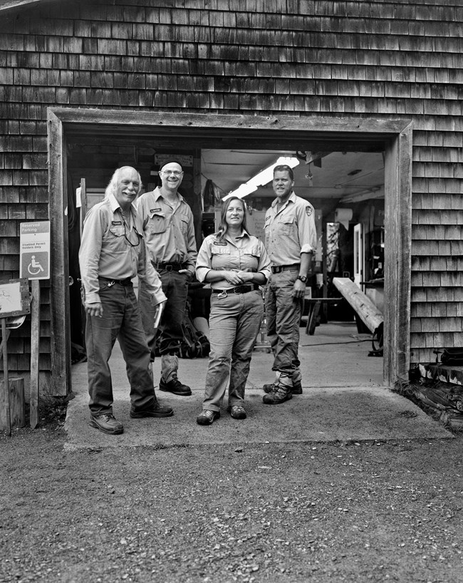 Black and white photo of four trail crew workers in front of the trails shop. All are smiling.
