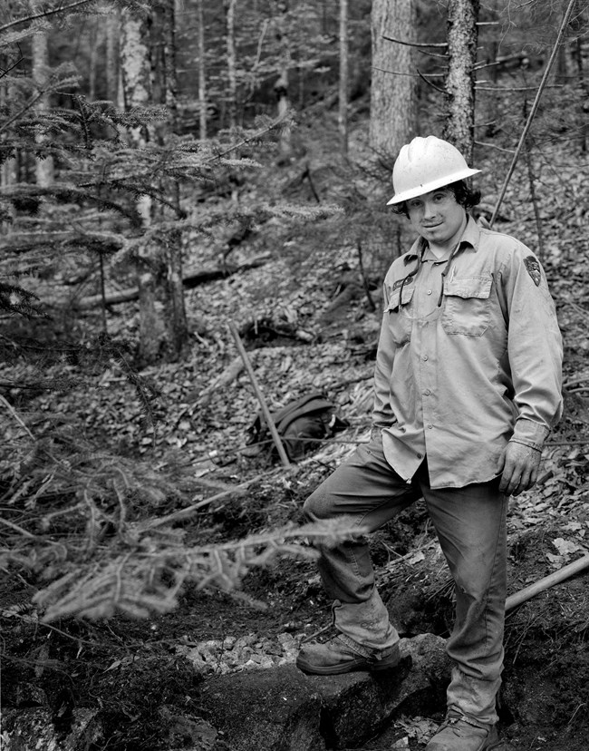 Black and white photograph of a male trail crew worker with a hard hat on and one leg stepping on a large rock.