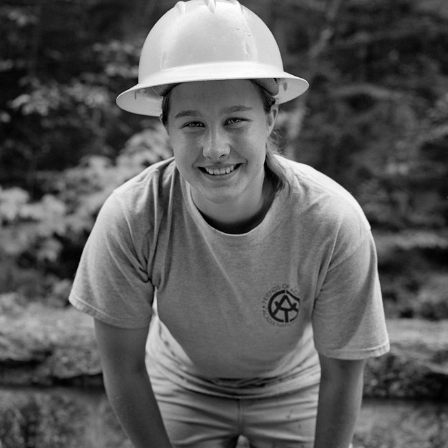 Black and white photo of young trail crew worker with her hat on. Forest scene in the background.