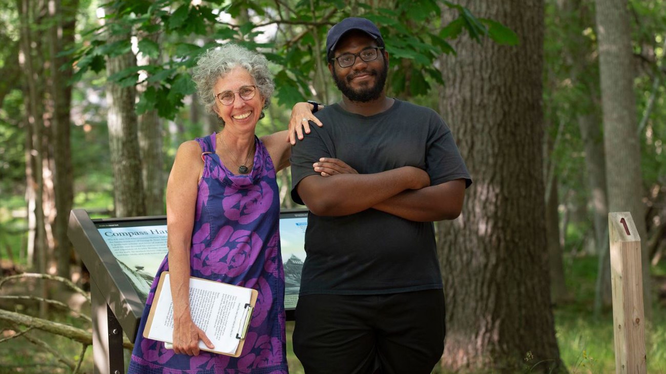 In a wooded area, a woman holding a clipboard rests her elbow on the shoulder of a man standing beside her