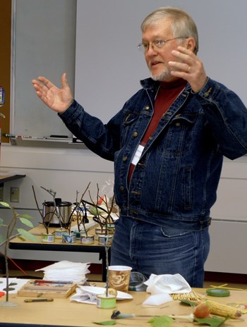 Man with glasses and denim jacket stands at table littered with art supplies