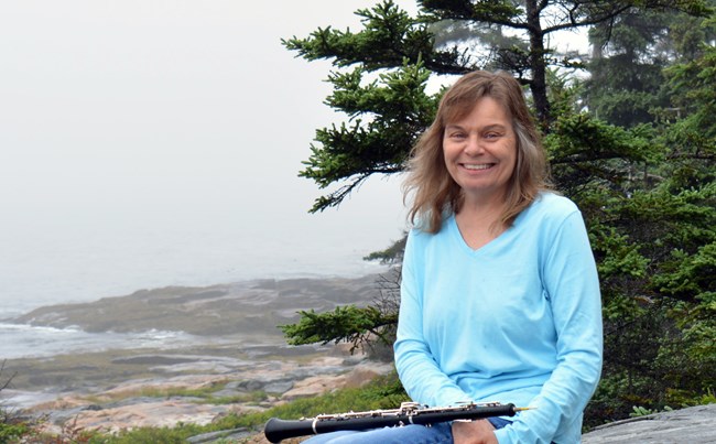 Woman with oboe on her lap smiles with trees and coastline in background Woman with oboe on her lap smiles with trees and coastline in background