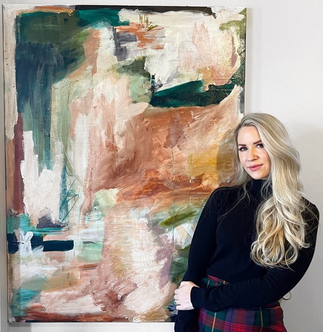 Kelly Collette with painting Woman with long blond hair and black sweater stands in front of a fine art painting on a wall