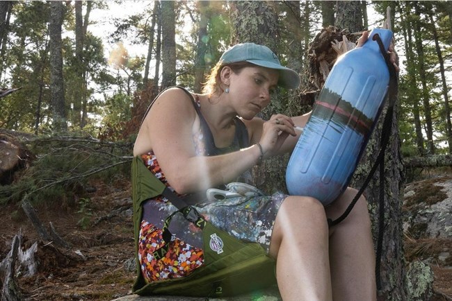 Woman sitting among trees paints a scene on a buoy