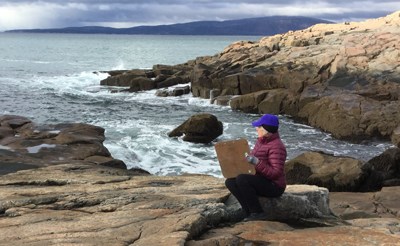 Woman bundled in winter clothes sits on rock to sketch ocean coastline