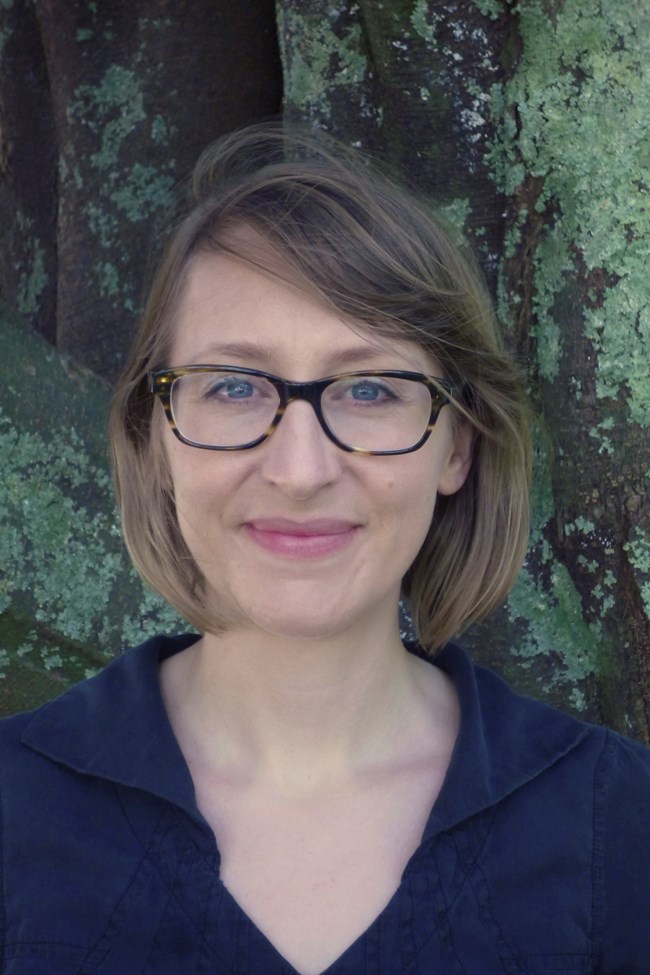 Portrait of a woman wearing glasses standing in front of dark rock face covered with green lichen