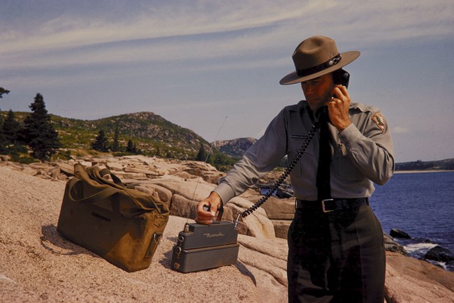A ranger at a rocky beach holds an early-model radio transmitter