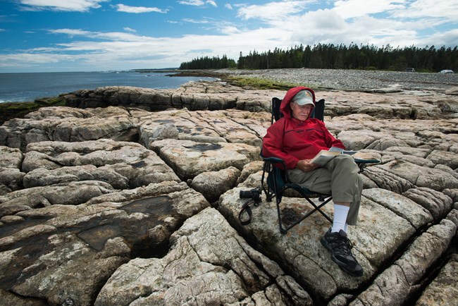 Man sitting in a packable trail chair reads a book on a stony outcropping along the ocean coastline.
