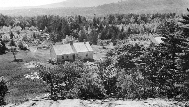 Historic black and white photograph of a simple small homestead house with mountains in the distance