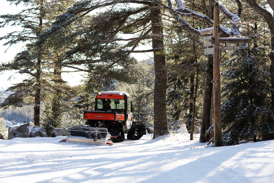 An orange, cabbed utility vehicle, drags grooming equipment behind it while driving on snow covered carriage roads in Acadia National Park on a sunny day. 