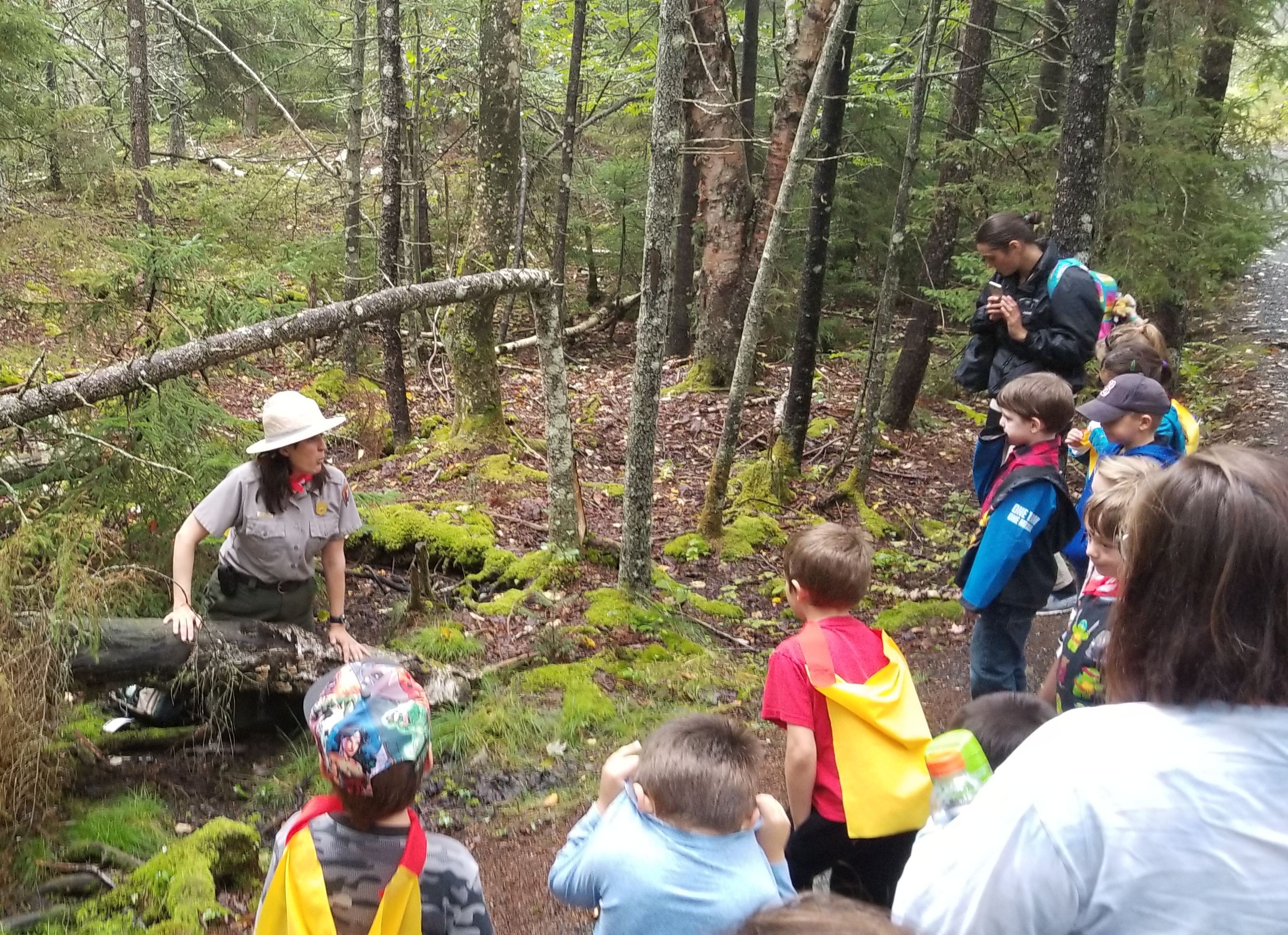 kids standing in capes and ranger kneels near fallen tree.